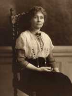 Formal portrait, Emmeline Pankhurst, three-quarter length, seated in chair, facing slightly right with head turned toward camera, wearing high-collared blouse with decorative buttons, bow tie, and necklace.
